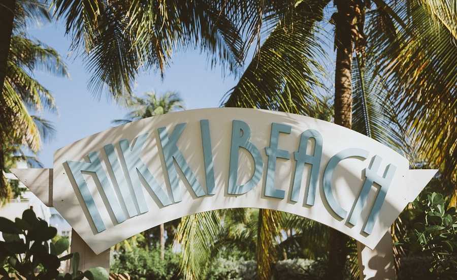 A sign that says "Miki Beach" in front of palm trees, located near nightclubs in Miami.