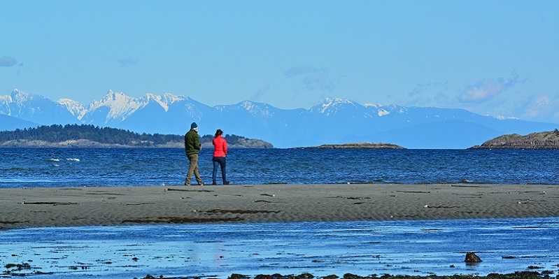 Two people standing on a Parksville beaches with mountains in the background.