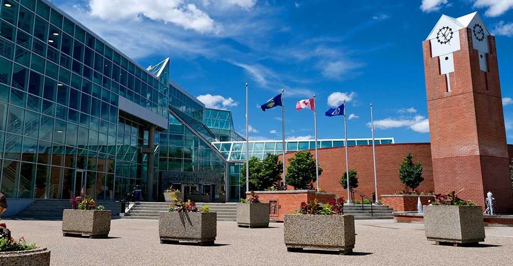 Modern public building with a glass facade and a clock tower, adorned with flower planters and flying flags under a blue sky with clouds, ranked among the best places to live in Alberta.