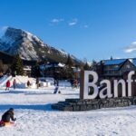 A winter scene in Banff, Canada, with people enjoying outdoor activities such as visiting the Banff Park Museum near a large "Banff" sign, with snowy mountains in the background.