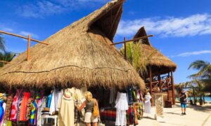 A woman shops at an outdoor market with thatched-roof stalls selling colorful clothing and accessories near a beach on a sunny day, reminiscent of vibrant scenes featured in any Mexico travel guide.