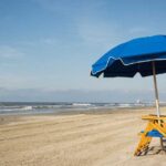 Galveston Island Beach boasts a sandy shore adorned with blue umbrellas and wooden chairs, all facing a calm ocean under a clear sky. It's easily one of the best beaches near Houston.