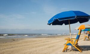 Galveston Island Beach boasts a sandy shore adorned with blue umbrellas and wooden chairs, all facing a calm ocean under a clear sky. It's easily one of the best beaches near Houston.