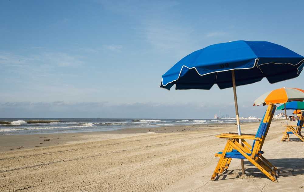 Galveston Island Beach boasts a sandy shore adorned with blue umbrellas and wooden chairs, all facing a calm ocean under a clear sky. It's easily one of the best beaches near Houston.