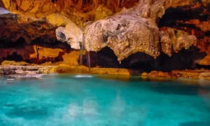 A stunning underground cave with turquoise water, a small waterfall, and stalactite formations hanging from the ceiling, reminiscent of the enchanting cave and basin hot springs in Banff National Park.
