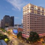 A cityscape at dusk featuring a tall, lit building in the foreground and a domed structure visible in the distance. Trees and streetlights line the road, with light traffic moving along, capturing the essence of downtown near some of the top 5-star hotels in Austin.