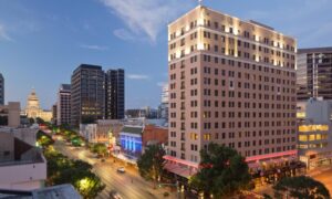 A cityscape at dusk featuring a tall, lit building in the foreground and a domed structure visible in the distance. Trees and streetlights line the road, with light traffic moving along, capturing the essence of downtown near some of the top 5-star hotels in Austin.