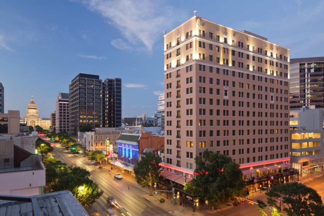 A cityscape at dusk featuring a tall, lit building in the foreground and a domed structure visible in the distance. Trees and streetlights line the road, with light traffic moving along, capturing the essence of downtown near some of the top 5-star hotels in Austin.