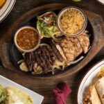 A table set with various dishes including a sizzling platter of grilled meats, a bowl of beans, guacamole, rice, a soda bottle, and a plate with a burrito and rice whispers of affordable places to eat in Houston.