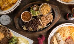 A table set with various dishes including a sizzling platter of grilled meats, a bowl of beans, guacamole, rice, a soda bottle, and a plate with a burrito and rice whispers of affordable places to eat in Houston.