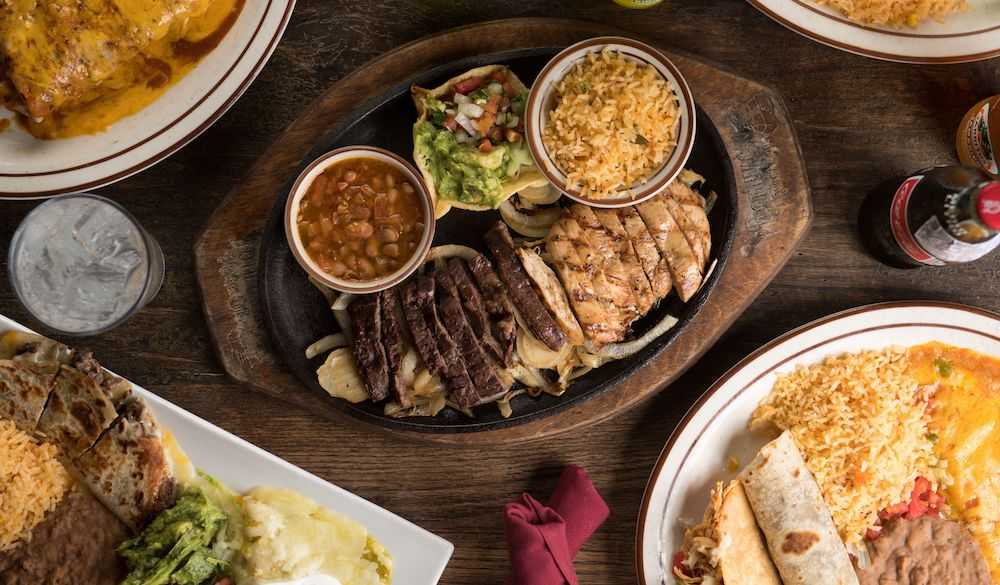 A table set with various dishes including a sizzling platter of grilled meats, a bowl of beans, guacamole, rice, a soda bottle, and a plate with a burrito and rice whispers of affordable places to eat in Houston.